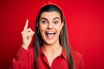 Wall Mural - Young beautiful brunette woman wearing casual shirt and diadem over red background pointing finger up with successful idea. Exited and happy. Number one.