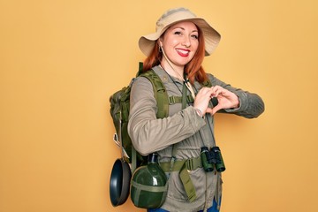 Poster - Young redhead backpacker woman hiking wearing backpack and hat over yellow background smiling in love doing heart symbol shape with hands. Romantic concept.