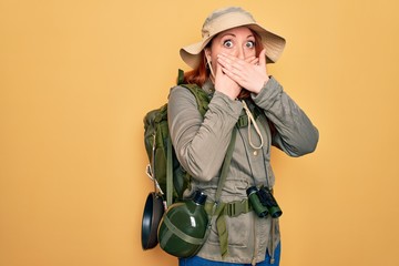 Poster - Young redhead backpacker woman hiking wearing backpack and hat over yellow background shocked covering mouth with hands for mistake. Secret concept.