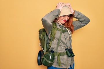 Poster - Young redhead backpacker woman hiking wearing backpack and hat over yellow background suffering from headache desperate and stressed because pain and migraine. Hands on head.