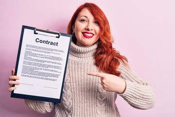 Sticker - Young redhead woman holding clipboard with contract document paper over pink background smiling happy pointing with hand and finger