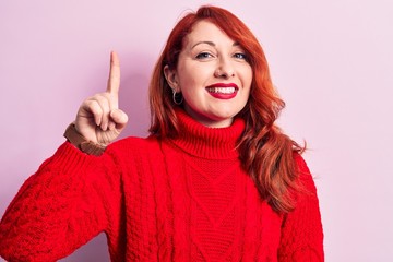 Poster - Young beautiful redhead woman wearing red casual turtleneck sweater over pink background smiling with an idea or question pointing finger up with happy face, number one