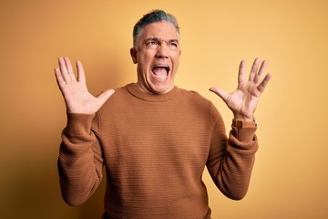 Poster - Middle age handsome grey-haired man wearing casual sweater over yellow background crazy and mad shouting and yelling with aggressive expression and arms raised. Frustration concept.