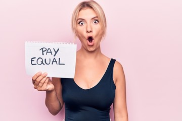 Beautiful blonde woman asking for equality economy holding paper with pay equal message scared and amazed with open mouth for surprise, disbelief face