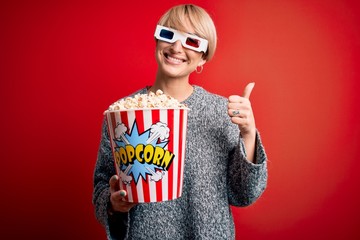 Poster - Young blonde woman with short hair wearing 3d movie glasses and eating popcorn at the cinema happy with big smile doing ok sign, thumb up with fingers, excellent sign