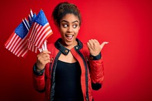 Young African American Patriotic Girl Holding United States Flags Over Isolated Red Background Pointing And Showing With Thumb Up To The Side With Happy Face Smiling