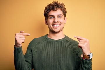 Young blond handsome man with curly hair wearing green sweater over yellow background looking confident with smile on face, pointing oneself with fingers proud and happy.