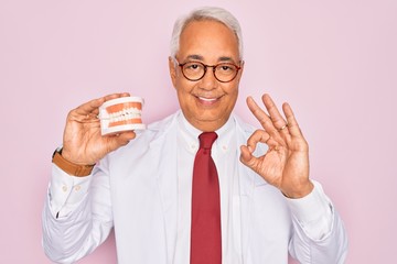 Wall Mural - Middle age senior grey-haired dentist man holding prosthesis denture over pink background doing ok sign with fingers, excellent symbol