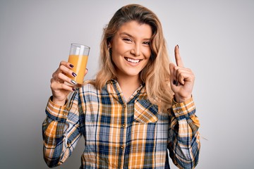 Poster - Young beautiful blonde woman drinking glass of healthy orange juice over white background surprised with an idea or question pointing finger with happy face, number one