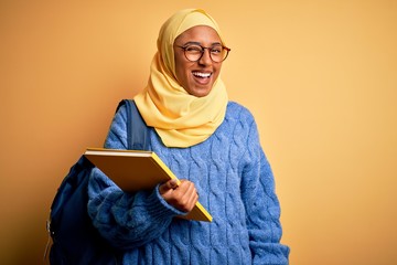 Wall Mural - Young African American student woman wearing muslim hijab and backpack holding book winking looking at the camera with sexy expression, cheerful and happy face.