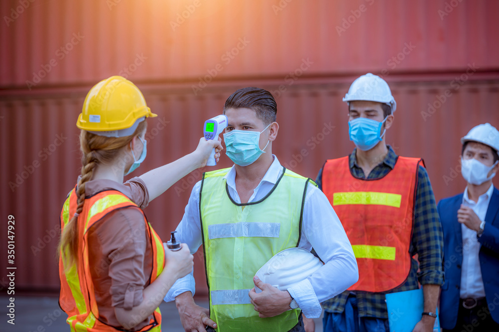 Woman worker used a medical Digital  temperature Thermometer to check staff body temperature before start work in factory ,they wearing face mask the concept of a corona virus [Covid-19] screening. - obrazy, fototapety, plakaty 