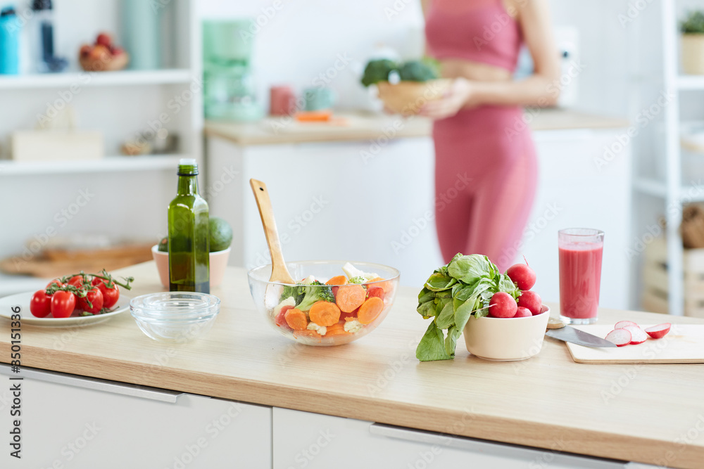 Close up of healthy food ingredients and salad bowl on kitchen counter with blurred shape of fit young woman in background, copy space - obrazy, fototapety, plakaty 