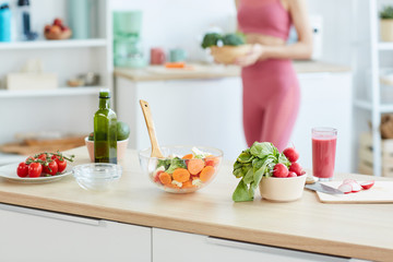 Close up of healthy food ingredients and salad bowl on kitchen counter with blurred shape of fit young woman in background, copy space