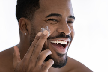 Close up portrait of happy young african American man apply moisturizing face cream for healthy glowing skin, smiling millennial biracial male use nourishing facial balm, skincare, hygiene concept