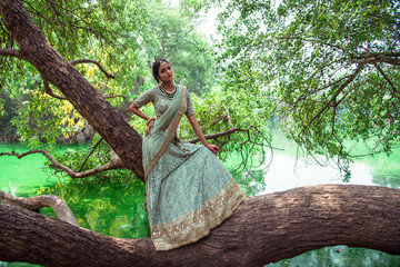 ull length portrait of an young beautiful Indian female model in traditional green designer dress saree in a park. Fashion photography on a tree in a summer park with a green river