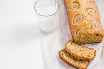 Wall Mural - Homemade sliced healthy banana bread ready for serve on breakfast on the white concrete background with craft baking paper and pastry knife, glass of milk. Top view. Close up shoot