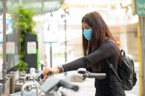 Cuadros en Lienzo Profile view of young Indian woman with mask riding bike at public bicycle servi