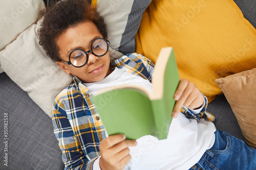 Cuadros en Lienzo Above view portrait of cute African boy wearing big glasses reading book while l