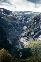 Wall Mural - Trip to Norway. Blue ice tongue of Jostedal glacier melts from the giant rocky mountains into the green valley with a lot of waterfalls. Vertical wallpaper