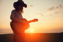 Silhouette Of Young Free Woman In Straw Hat Playing Country Music On A Guitar At Sunset, Copy Space