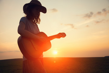 silhouette of young free woman in straw hat playing country music on a guitar at sunset, copy space