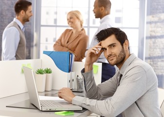 Wall Mural - Portrait of young businessman sitting at desk, having laptop, working, looking at camera.