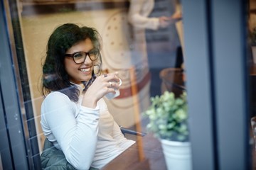 Poster - Happy woman sitting in cafeteria, drinking coffee, smiling. Photographed through window.