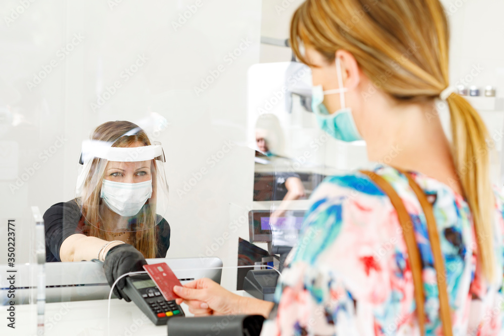 dentro un locale commerciale la cliente paga alla cassa con carta di credito munita di mascherina e separata dalla cassiera da un separatore di plexiglass  - obrazy, fototapety, plakaty 