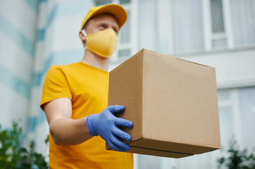 Delivery Man worker in yellow uniform cap, t-shirt, face mask and gloves holds a cardboard box package on building backdrop. Safety delivery quarantine service in covid-19 virus pandemic period.