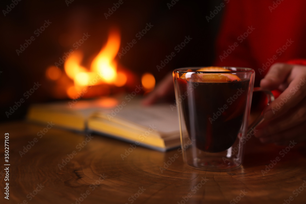 Woman with mulled wine reading book near fireplace indoors, closeup. Winter vacation - obrazy, fototapety, plakaty 