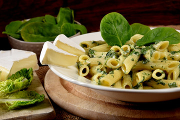 Wall Mural - penne in spinach sauce on a white plate in the company of fresh spinach leaves and blue cheese on a wooden kitchen board