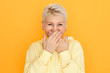 © Anatoliy Karlyuk - Studio shot of cheerful beautiful European female pensioner with dyed pixie hairstyle covering her mouth with both hands while giggling, laughing at joke, having shy look, supressing laughter
