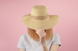 Redhair girl in gray tshirt hiding behind a straw hat as she pulls it down over her face with her hands. Frontal view upper body pink