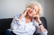 © mimagephotos - elderly woman sitting on sofa with headache and hands to head