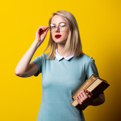 Wall Mural - Blonde girl in blue dress and glasses with books on yellow background