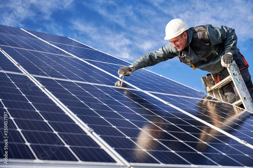 Side view of joyful male technician in safety helmet standing on ladder and installing photovoltaic solar panel under beautiful blue sky. Electrician in gloves mounting stand-alone solar panel system.