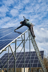 Wall Mural - Vertical snapshot of working process in a solar field. Technician in protective uniform, standing on a ladder checking solar batteries on metal ground mounting. Concept of renewable energy resource