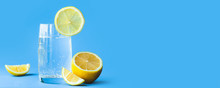 Translucent Glass Of Water With Gases And Bubbles, With A Round Slice Of Lemon On A Glass And A Whole Lemon On Blue Background. Refreshment In The Heat. Healthy Banner Lifestyle And Nutrition