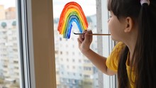 The Child, Beautiful Girl Draws A Rainbow On The Window. Stay Home, Flashmob Chase The Rainbow. Covid-19 Quarantine, Coronavirus Pandemic. Happy Child Shows Pozitiv Emotions, Paint On The Window Glass