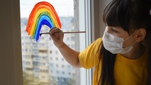 The Child, Little Girl In The Medical Mask, Smiles Draws A Rainbow On The Window. Stay Home, Flashmob Chase The Rainbow. Covid-19 Quarantine, Coronavirus. Happy Child, Pozitiv Emotions And Painting