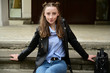 Model posing sitting in spring park outdoors in the city. Photo of a young pretty girl with a smile in a black jacket and jeans.