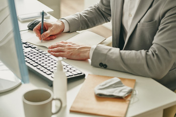 Poster - Close-up of businessman sitting at desk with sanitizer and mask and making notes in notepad while working in office during coronavirus quarantine