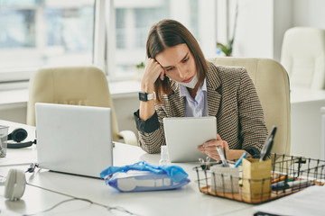Poster - Serious tired young businesswoman with mask on chin sitting at desk with laptop and checking mail on tablet in empty office during covid-19