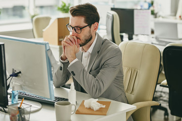 Wall Mural - Nervous young businessman in eyeglasses sitting with closed eyes and trying to focus on work in empty office during coronavirus