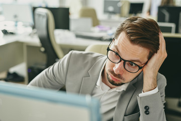 Wall Mural - Stressed young businessman in glasses working alone in office and looking at bad sales statistics on computer monitor during coronavirus pandemic