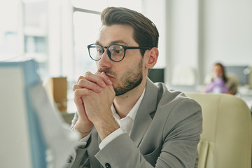 Poster - Concentrated young bearded businessman leaning on hands and looking at computer monitor while praying about concluding deal during coronavirus