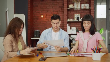 Wall Mural - Creative startup business team of three discussing printouts with website interface templates while planning project sitting at wooden desk in loft office