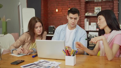Wall Mural - Creative young business team of three looking at laptop screen, discussing ideas and planning project together sitting at wooden desk in modern loft office