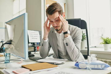 Wall Mural - Confused young businessman in jacket sitting at desk in empty office and massaging temples while reading agreement