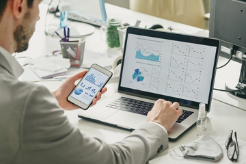 Wall Mural - Over shoulder view of businessman using smartphone and laptop while analyzing sales graphs during quarantine